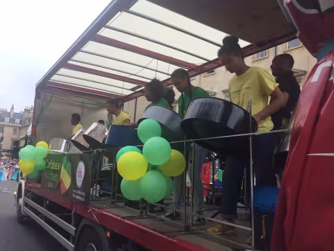 Young people on a truck serving as a float, playing steel pan, dressed in green and yellow with balloons at a carnival event.